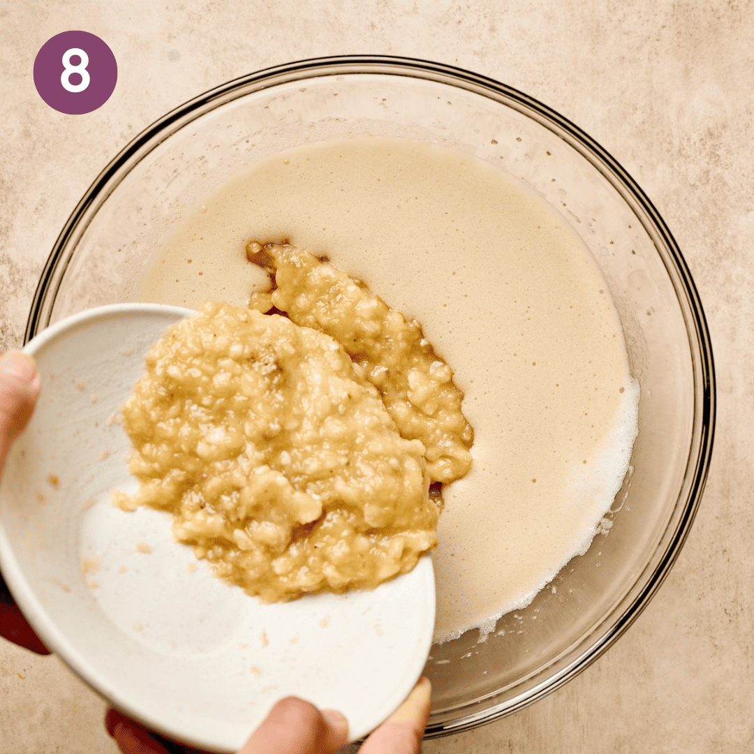 Person adding a bowl of mashed bananas to the mixture of wet ingredients.