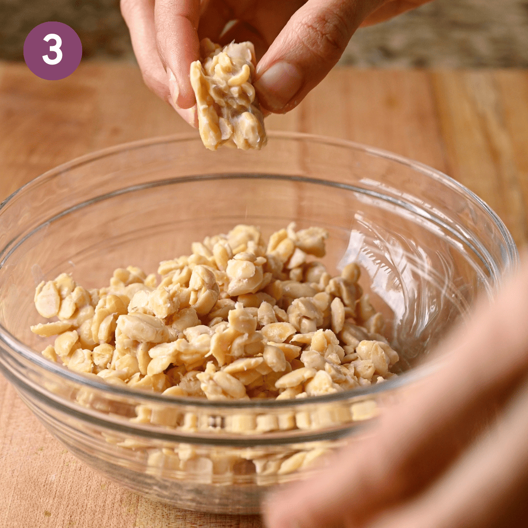 person crumbling tempeh into a glass bowl on a wooden cutting board.