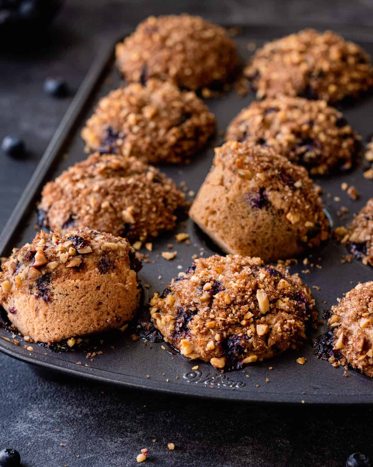 blueberry muffins with crumble topping in muffin tin, two muffins spilling out.