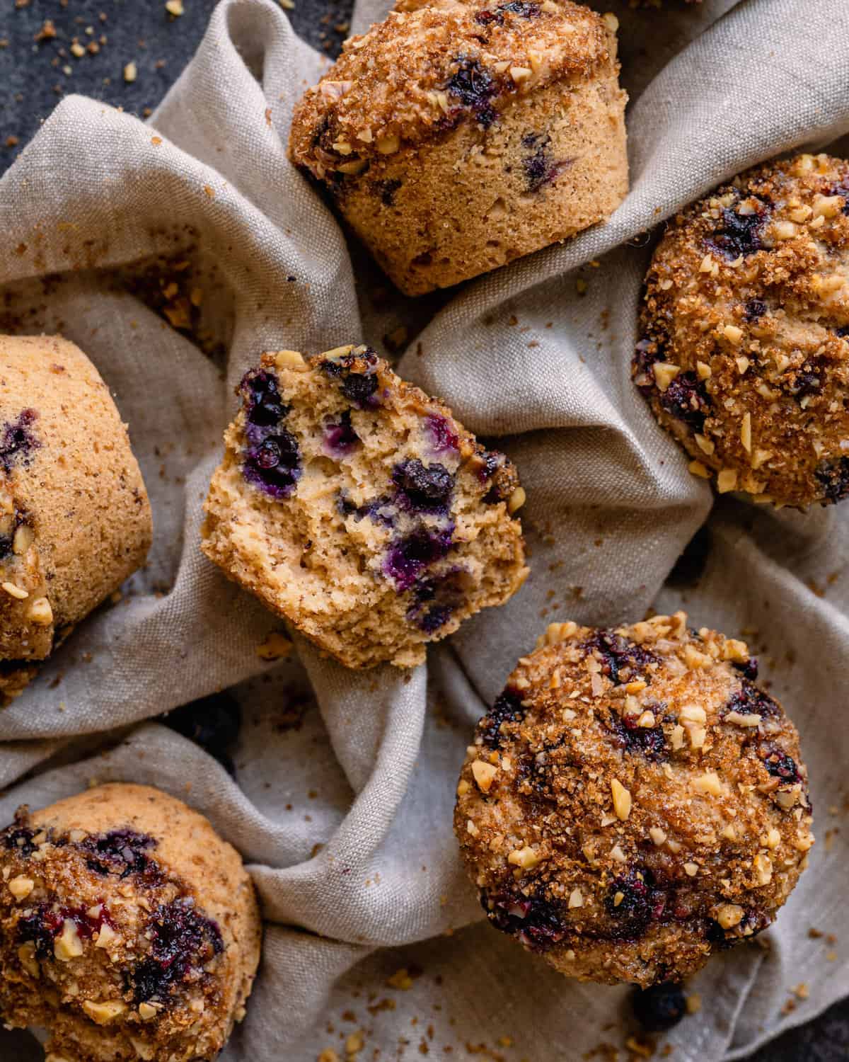 flatlay of vegan blueberry muffins on linen with middle muffin sliced open to show texture.