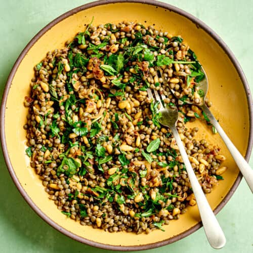 Spoon and fork in lentil salad in a yellow bowl on green table.