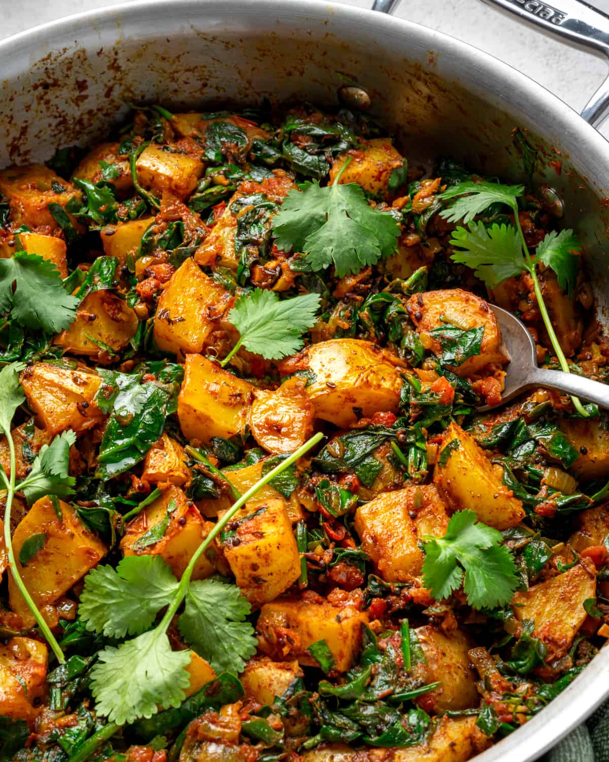close up photo of saag aloo in saucepan with cilantro leaves and a spoon.