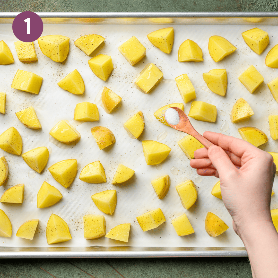 person sprinkling salt over cubed potatoes on a lined baking tray.