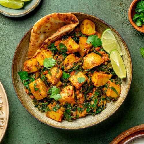 bowl of saag aloo with naan and lime wedges on a table.