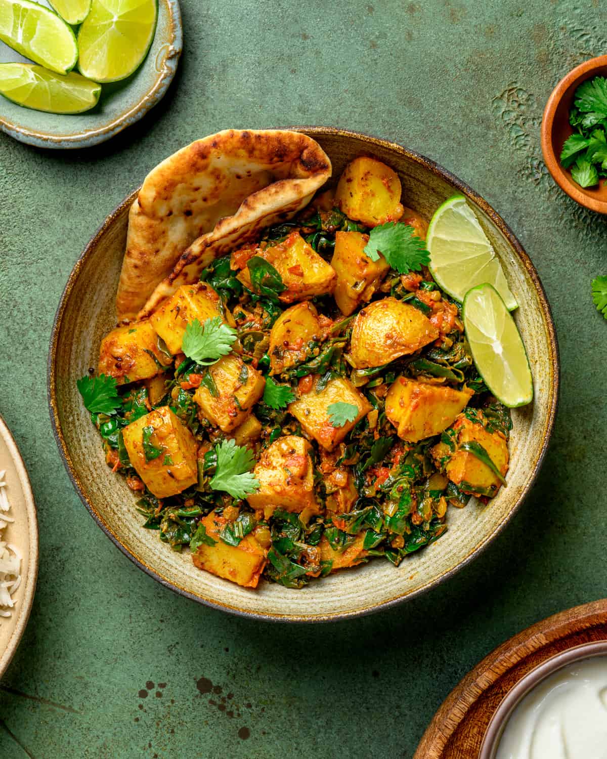 bowl of saag aloo with naan and lime wedges on a dark green table.