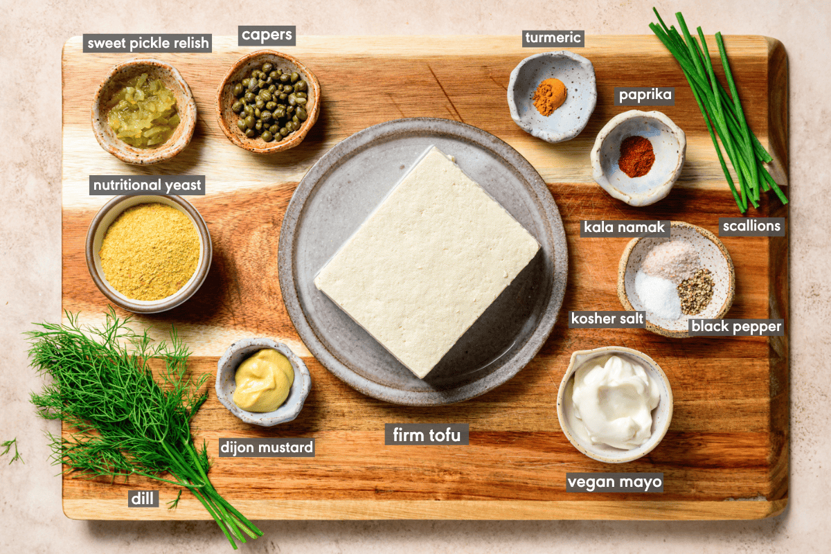 Vegan egg salad ingredients in various small bowls on a wooden cutting board.